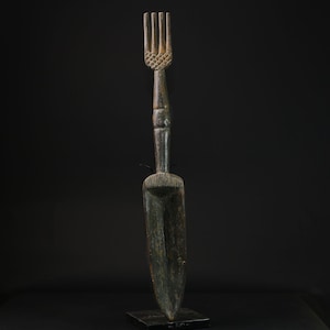 Op de afbeelding: Een hoge, donkere houten sculptuur met een vorkachtig bovenstuk en een lange, slanke steel. De vork heeft drie tanden en een gestructureerd ontwerp. De steel leidt naar een brede, platte, lepelvormige basis. De sculptuur is gemonteerd op een kleine, vierkante basis.