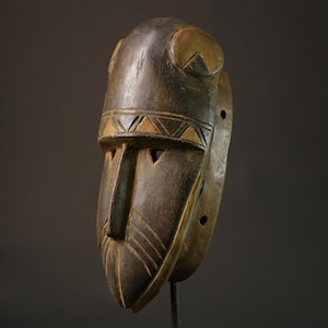 Puede incluir: Una máscara de madera con una cara tallada y patrones geométricos. La máscara es de color marrón oscuro con acentos marrones claros. Tiene una forma redondeada y está montada en un soporte negro.