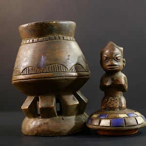 Puede incluir: Una escultura de madera tallada con una base grande y bulbosa y una figura separada. La base tiene patrones geométricos tallados. La figura es una persona pequeña y sentada con un rostro detallado, sobre una base con detalles azules y blancos.