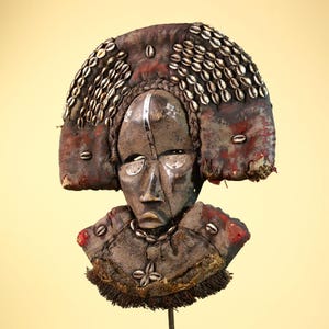 Könnte beinhalten: Eine verwitterte Stammesmaske mit einem dunkelbraunen Gesicht und einem Kopfschmuck, der mit zahlreichen kleinen, weißen Muscheln verziert ist. Die Maske hat eine markante Nase, Augenausschnitte und einen dekorativen Kragen. Die Farbpalette ist erdig, mit Braun- und Rottönen.