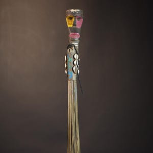 African traditional fly whisk with long plant fiber tassels, a carved wooden handle featuring a painted tribal face, and cowrie shell accents. Handcrafted ceremonial whip tool used in African rituals, displayed against a dark background. Vintage wall art or decor piece reflecting authentic African heritage and craftsmanship.
