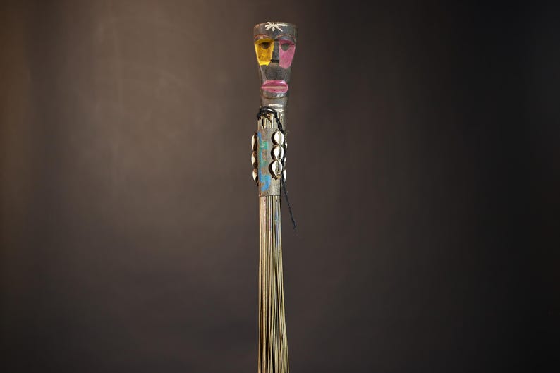 African traditional fly whisk with long plant fiber tassels, a carved wooden handle featuring a painted tribal face, and cowrie shell accents. Handcrafted ceremonial whip tool used in African rituals, displayed against a dark background. Vintage wall art or decor piece reflecting authentic African heritage and craftsmanship.