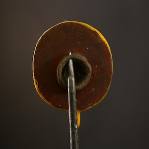 Op de afbeelding: Close-up van de achterkant van een rond, geel en rood geschilderd houten ornament. Het ornament heeft een metalen staaf die uit het midden steekt.