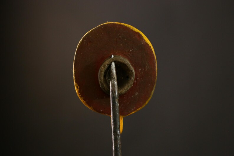 Op de afbeelding: Close-up van de achterkant van een rond, geel en rood geschilderd houten ornament. Het ornament heeft een metalen staaf die uit het midden steekt.