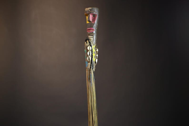 African traditional fly whisk with long plant fiber tassels, a carved wooden handle featuring a painted tribal face, and cowrie shell accents. Handcrafted ceremonial whip tool used in African rituals, displayed against a dark background. Vintage wall art or decor piece reflecting authentic African heritage and craftsmanship.