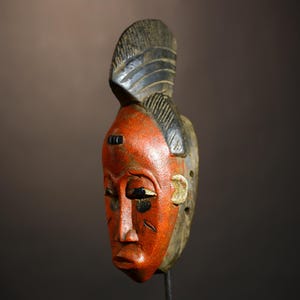 May include: A wooden mask with a red face and black details. The mask has a tall, black, carved headdress.