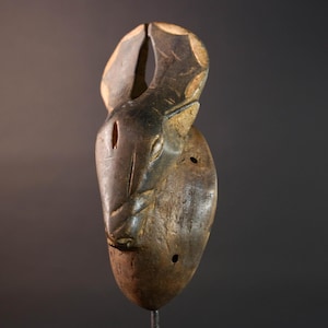 Puede incluir: Una máscara de madera tallada con un acabado marrón oscuro. La máscara presenta una cara de animal estilizada con una forma ovalada larga y un diseño prominente de boca abierta. La máscara tiene una parte superior grande y curvada y dos pequeños agujeros a los lados.