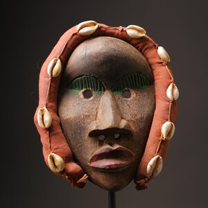 Könnte beinhalten: Eine Holzmaske mit einem menschlichen Gesicht, bemalt in Braun, Grün und Rot. Die Maske ist mit weißen Kaurischneckenmuscheln an den Rändern verziert.