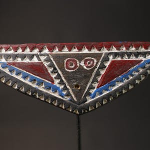 Op de afbeelding: Een houten, driehoekig masker met een donkerbruine basis. Het masker heeft rood en blauw geschilderde driehoeken, witte accenten en een centrale driehoek met twee cirkelvormige oogontwerpen. De randen zijn afgezet met witte driehoeken.
