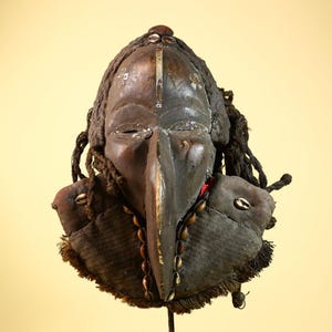 Könnte beinhalten: Eine geschnitzte Holzmaske mit einer langen, schnabelartigen Nase und aufwendigen Details. Die Maske ist überwiegend braun mit dunkleren Akzenten, verziert mit Muscheln und seilartigen Elementen. Sie zeigt ein stilisiertes Gesicht und ist wahrscheinlich von kultureller Bedeutung.