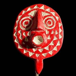 May include: A red and white ceramic mask with a stylized face and a circular design. The mask has a large, open mouth and two large eyes with white circles around them.