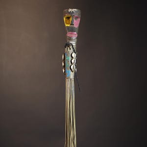 African traditional fly whisk with long plant fiber tassels, a carved wooden handle featuring a painted tribal face, and cowrie shell accents. Handcrafted ceremonial whip tool used in African rituals, displayed against a dark background. Vintage wall art or decor piece reflecting authentic African heritage and craftsmanship.