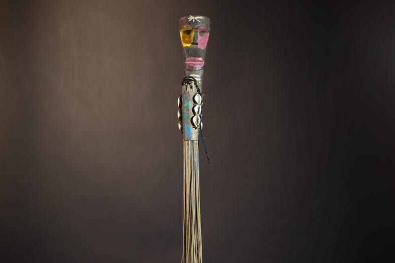 African traditional fly whisk with long plant fiber tassels, a carved wooden handle featuring a painted tribal face, and cowrie shell accents. Handcrafted ceremonial whip tool used in African rituals, displayed against a dark background. Vintage wall art or decor piece reflecting authentic African heritage and craftsmanship.