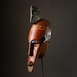 May include: A wooden mask with a red, blue, and yellow paint design. The mask has a long, narrow face with a pointed chin and a small, round mouth. The eyes are large and almond-shaped, and the nose is long and thin. The mask is decorated with a variety of geometric patterns and symbols.