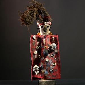 May include: A handcrafted wooden doll with a red cloth cloak decorated with shells and beads. The doll has a carved face, a headdress, and a bundle of dark fibres extending from the head. The doll is mounted on a wooden base, showcasing a unique, cultural aesthetic.
