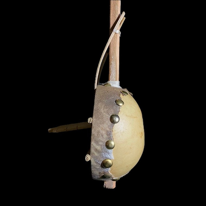 May include: A small, hand-crafted drum made from a gourd, with a leather head and brass tacks. The drum is attached to a wooden stick with a leather strap.