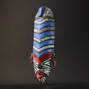 Könnte beinhalten: Eine Holzmaske mit einem blau-weiß-schwarzen Streifenmuster. Die Maske hat ein rotes und weißes Gesicht mit einem schwarz-weißen Zebra-Muster.