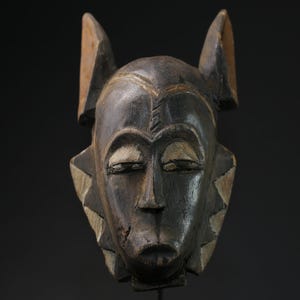 May include: A wooden mask with a dark brown finish. The mask has a human-like face with closed eyes, a prominent nose, and a slightly open mouth. The mask has two large, pointed ears that extend upwards.