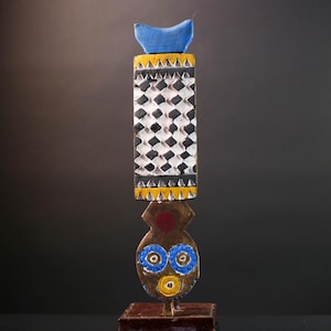 May include: A tall, handcrafted wooden sculpture with a geometric design. The sculpture features a blue top, a checkered black and white middle section, and a face-like lower portion with blue and yellow details. It stands on a small, dark brown base.