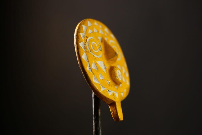 Op de afbeelding: Een gele zonvormige ornament met een wit gezicht en geometrische ontwerpen. De ornament is op een zwarte achtergrond.