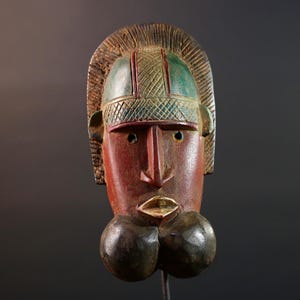 May include: A carved wooden mask with a red face, green and brown headdress, and dark brown lower section. The mask has a prominent nose, open mouth, and two bulbous shapes below the mouth. The mask is displayed against a dark background.