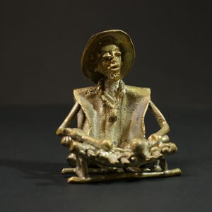 Peut inclure: Une sculpture en métal de couleur dorée représentant une personne portant un chapeau et assise sur une chaise. La personne tient un instrument de musique sur ses genoux.