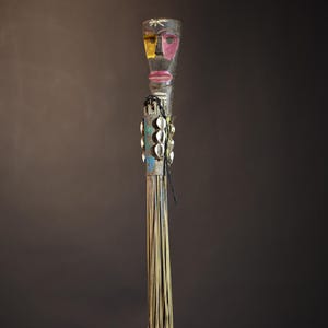 African traditional fly whisk with long plant fiber tassels, a carved wooden handle featuring a painted tribal face, and cowrie shell accents. Handcrafted ceremonial whip tool used in African rituals, displayed against a dark background. Vintage wall art or decor piece reflecting authentic African heritage and craftsmanship.