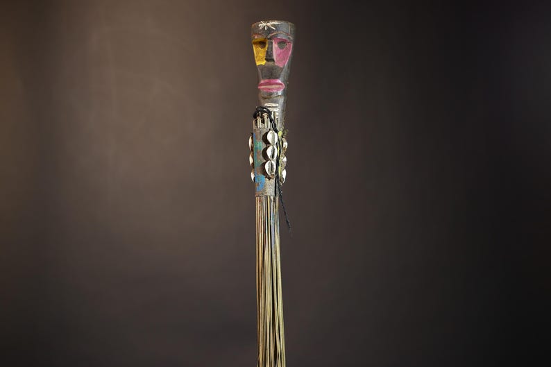 African traditional fly whisk with long plant fiber tassels, a carved wooden handle featuring a painted tribal face, and cowrie shell accents. Handcrafted ceremonial whip tool used in African rituals, displayed against a dark background. Vintage wall art or decor piece reflecting authentic African heritage and craftsmanship.