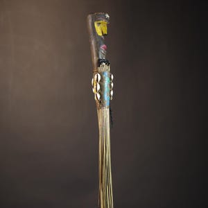 African traditional fly whisk with long plant fiber tassels, a carved wooden handle featuring a painted tribal face, and cowrie shell accents. Handcrafted ceremonial whip tool used in African rituals, displayed against a dark background. Vintage wall art or decor piece reflecting authentic African heritage and craftsmanship.