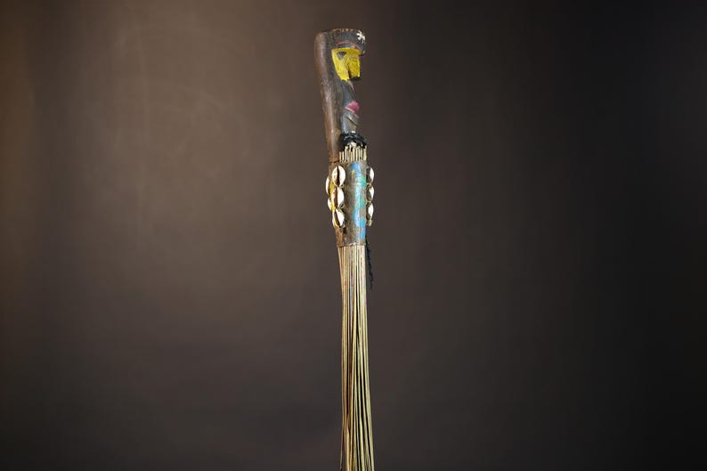 African traditional fly whisk with long plant fiber tassels, a carved wooden handle featuring a painted tribal face, and cowrie shell accents. Handcrafted ceremonial whip tool used in African rituals, displayed against a dark background. Vintage wall art or decor piece reflecting authentic African heritage and craftsmanship.