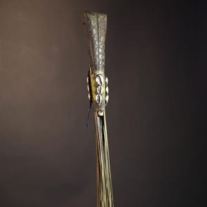 African traditional fly whisk with long plant fiber tassels, a carved wooden handle featuring a painted tribal face, and cowrie shell accents. Handcrafted ceremonial whip tool used in African rituals, displayed against a dark background. Vintage wall art or decor piece reflecting authentic African heritage and craftsmanship.