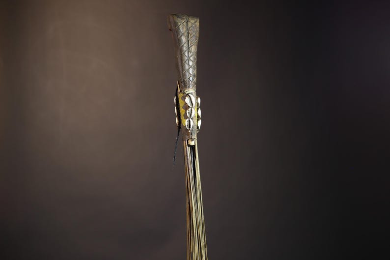 African traditional fly whisk with long plant fiber tassels, a carved wooden handle featuring a painted tribal face, and cowrie shell accents. Handcrafted ceremonial whip tool used in African rituals, displayed against a dark background. Vintage wall art or decor piece reflecting authentic African heritage and craftsmanship.