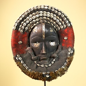 Könnte beinhalten: Eine runde, dunkle Holzmaske mit geschnitztem Gesicht, verziert mit weißen Muscheln und einem rot-schwarzen Rand. Die Maske hat Metallakzente und einen pelzartigen Besatz, die einen Stammeskunst-Stil zeigen.