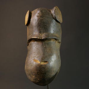 Puede incluir: Una máscara de madera marrón oscuro con un rostro humano. La máscara tiene una nariz ancha y plana y ojos grandes y redondos. La máscara está tallada con intrincados detalles y tiene un acabado liso y pulido.