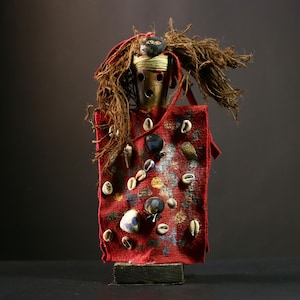May include: A red fabric doll with brown hair-like material and a wooden face. The doll is decorated with white and blue shells and other embellishments.