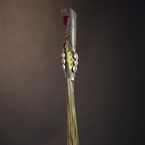 African traditional fly whisk with long plant fiber tassels, a carved wooden handle featuring a painted tribal face, and cowrie shell accents. Handcrafted ceremonial whip tool used in African rituals, displayed against a dark background. Vintage wall art or decor piece reflecting authentic African heritage and craftsmanship.