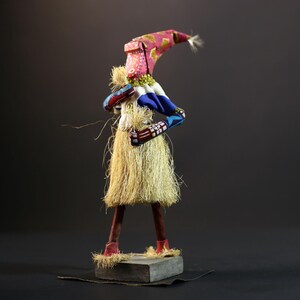 May include: A colorful handmade doll with a pink and gold hat, blue and white patterned fabric, and a long, straw-like skirt. The doll is standing on a wooden base.