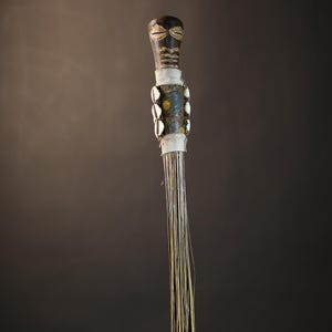 May include: A wooden ceremonial staff with a carved face at the top. The staff is decorated with white shells and a white cloth. The staff is made of natural materials and has a rustic appearance.