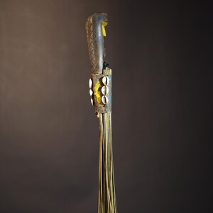 African traditional fly whisk with long plant fiber tassels, a carved wooden handle featuring a painted tribal face, and cowrie shell accents. Handcrafted ceremonial whip tool used in African rituals, displayed against a dark background. Vintage wall art or decor piece reflecting authentic African heritage and craftsmanship.