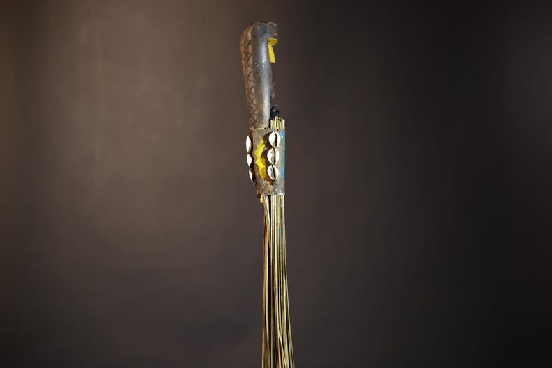African traditional fly whisk with long plant fiber tassels, a carved wooden handle featuring a painted tribal face, and cowrie shell accents. Handcrafted ceremonial whip tool used in African rituals, displayed against a dark background. Vintage wall art or decor piece reflecting authentic African heritage and craftsmanship.