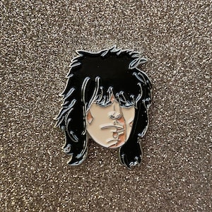 May include: A black and white enamel pin featuring a cartoon portrait of a person with black hair and a white face. The person has a black eye and a red mark on their cheek.