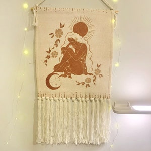 May include: A woven wall tapestry with a brown silhouette of a person sitting on a crescent moon with a sun above. The tapestry has a fringe border and is decorated with floral designs.