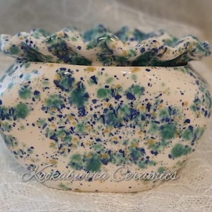 May include: A ceramic bowl with a frilled edge, decorated with a speckled pattern of blue, green, and yellow on a white background. The bowl has a rounded shape and the text "Kookaburra Ceramics" is visible on the front.