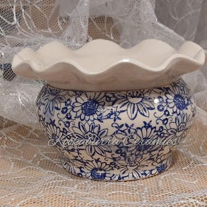 May include: A ceramic bowl with a ruffled rim and a blue floral pattern on a cream background. The text Kookaburra Ceramics is printed in a cursive script across the front of the bowl. The bowl has a rounded body and a smooth, glossy finish.