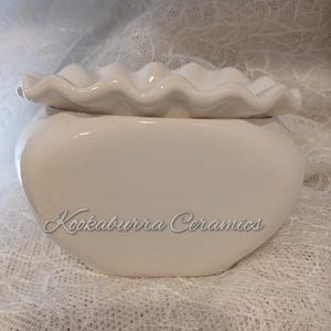 May include: A white ceramic pot with a ruffled lid. The pot has a rounded shape and the text "Kookaburra Ceramics" is written on the front. The lid has a wavy, scalloped edge.