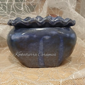 May include: A dark blue ceramic planter with a ruffled, wavy rim and a smooth, glossy finish. The body of the planter features vertical drip-like glaze patterns in lighter blue tones. The text Kookaburra Ceramics is printed in white on the front.