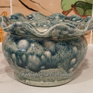 May include: A ceramic bowl with a ruffled edge and a speckled blue glaze. The bowl has a round base and a lid. The text "Kookaburra Ceramics" is written on the side of the bowl.
