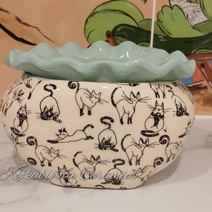 May include: A ceramic bowl with a light blue, frilled rim and a cream base. The base is decorated with black cat illustrations in various poses. The bowl is likely handmade and suitable for home decor.
