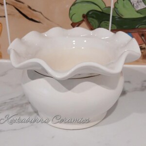 May include: A white ceramic bowl with a frilled edge, set on a rounded base. The bowl has a smooth, glossy finish. The text "Kookaburra Ceramics" is written in cursive on the base.