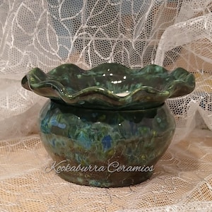 May include: A round ceramic bowl with a ruffled, wavy rim, featuring a glossy glaze in mottled shades of deep forest green, teal, and brown. The text Kookaburra Ceramics is printed in a white script font across the front of the bowl.